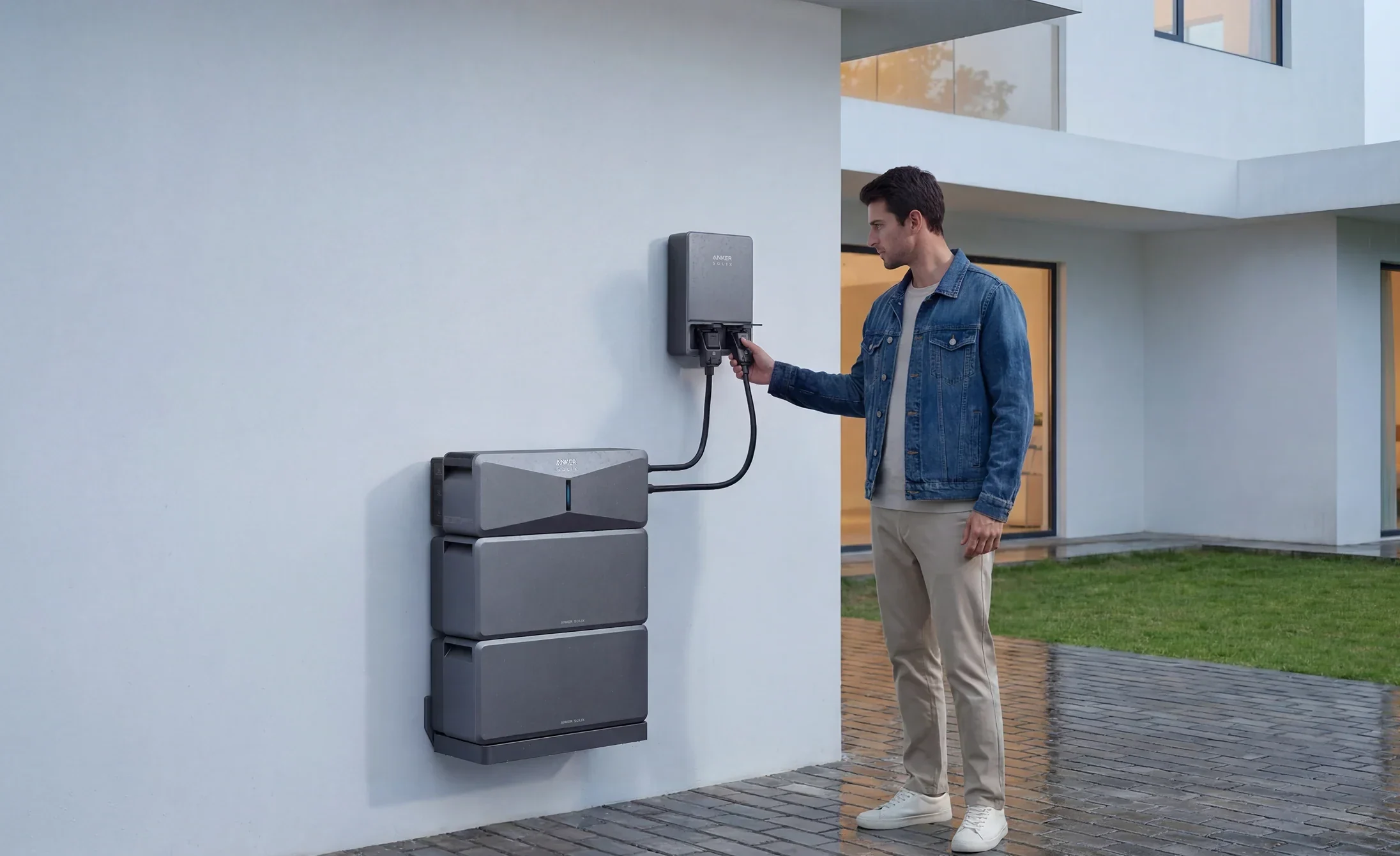 A man plugs a cable into an Anker SOLIX E10 home backup system mounted on an exterior house wall, the power conversion unit above three stacked battery modules.